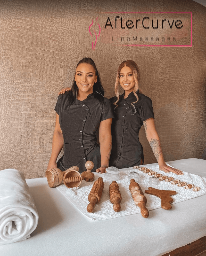 Angele en Kelly van AfterCurve bij de behandeltafel met houten massage-instrumenten, met het logo aan de wand.